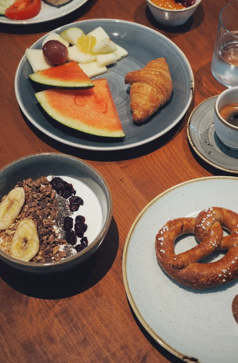 Eine Auswahl an Frühstücksspeisen, darunter Brezel, Obst, Croissant und Joghurt mit Müsli.